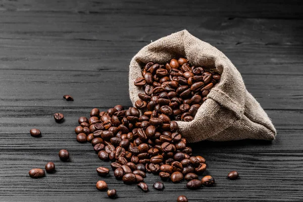Friss pörkölt kávé bab esik ki a zsákot, a fa felületén. Barna szemes kávé szétszórt táskát az asztalra — Stock Fotó