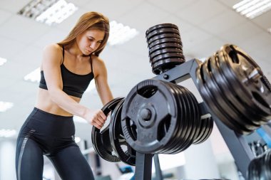 Siyah sportif giyim konsantre genç kadın spor salonunda barbell disk alarak. Güzel fitness bayan sağlıklı egzersiz sonrası özel standı içine barbell plaka koyar.