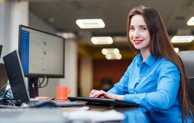 Başarılı kadın programcı bir bilgisayar ile masada oturuyor ve çalışır. Güzel kadın kamera bakıyor ve bir yazılım şirketi ofisinde gülümseyen.