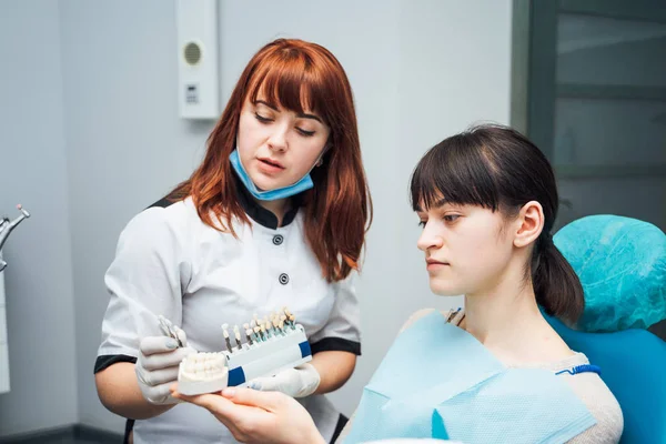 Die attraktive Zahnärztin zeigt einem Patientin, die auf dem Zahnarztstuhl sitzt, das Kieferlayout. Zahnarzt erklärt einer Patientin die Zahnfarben am Zahnmodell in der Stomatologie — Stockfoto