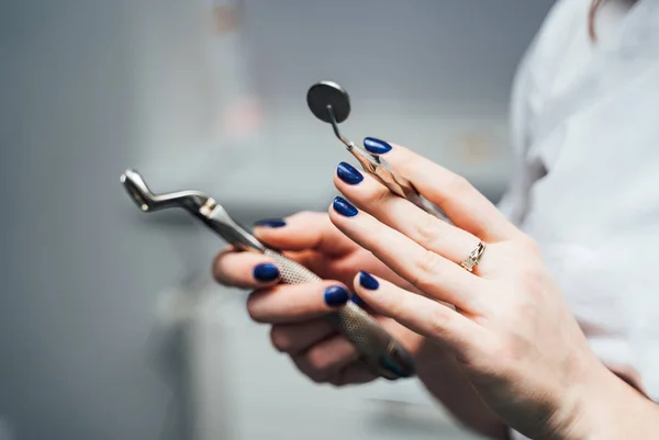 Hands of a stomatologist with blue nails are holding stainles instruments in clinic. Medical equipment in the hands of a dentist on the blurred background. Close-up. — Stock Photo, Image