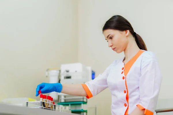 Tıp üniformalı güzel laboratuvar teknisyeni kadın kan örnekleriyle çalışıyor. Kadın araştırmacı laboratuvarda bir sürü sıvı şişesiyle çalışıyor.. — Stok fotoğraf