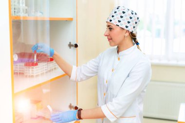 Tıbbi üniformalı bir kadın hemşirenin rafa test tüpleri koyarak yan görünümü. Beyaz laboratuvar önlüğü, şapka ve lateks eldiven giymiş profesyonel hemşire tıbbi bir laboratuvarda kan örnekleri yerleştirerek.