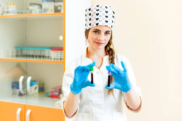 Laboratuvarda kanla iki test tüpü tutan büyüleyici bir sağlık çalışanının portresi. Tıbbi üniforma ve mavi steril eldiven kibar hemşire sıvı şişeleri gösterir ve kameraya bakar. — Stok fotoğraf
