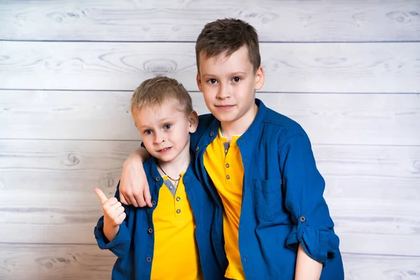 Twee aantrekkelijke jongens in blauwe shirts en gele t-shirts poseren samen in Studio. Portret van twee broers knuffelen elkaar en kijken naar de camera geïsoleerd op lichte houten achtergrond. — Stockfoto