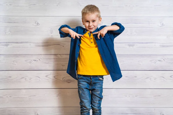 Menino emocional de camisa azul e jeans em pé no fundo da parede do estúdio de madeira. Criança loira feliz mostrando os dedos na frente dele e olhando para a câmera . — Fotografia de Stock