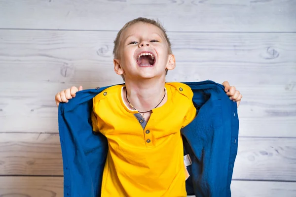 Retrato de meio comprimento de um menino alegre tirando sua camisa azul enquanto olha para a câmera. Menino feliz com a boca aberta em pé no fundo do estúdio de madeira luz . — Fotografia de Stock