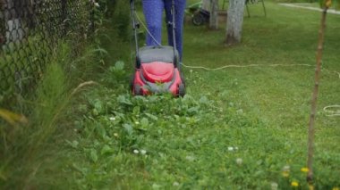 Bir kadın çitin yanında çim biçme makinesiyle çim biçiyor.