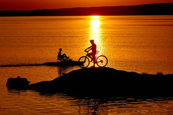 Mężczyzna rower jeździec stoi obok jego rower w pobliżu rzeki o zachodzie słońca. Sylwetka rowerzysty w kasku, który ma odpocząć na pomarańczowym tle wody. Wieczorny krajobraz i rowerzysta. — Zdjęcie stockowe