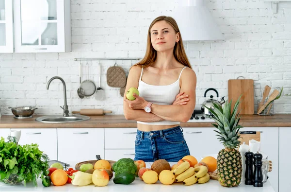 Jovem mulher de pé perto da mesa na cozinha, sorrindo, olhando para a câmera. Alimentação saudável e conceito de dieta . — Fotografia de Stock