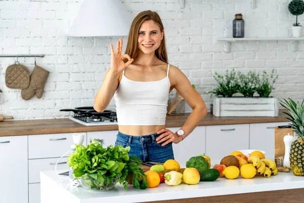 Mujer joven de pie en la cocina rodeada de verduras frescas mezcladas, sonriendo, mirando a la cámara . — Foto de Stock