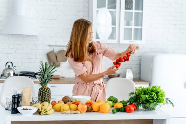 Mutfakta sebze ve meyvelerle yemek hazırlamaya hazır bir kadın var. Kadın elindeki domateslere bakıyor. Mutfak arka planı. Sağlıklı yiyecekler. Vejetaryen. Vejetaryen..