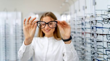 Mutlu kadın optometrist, optik dükkanında arka planda gözlük setiyle ayakta duruyor. Kadın gözlükleri elinde tutuyor. Yakın çekim..