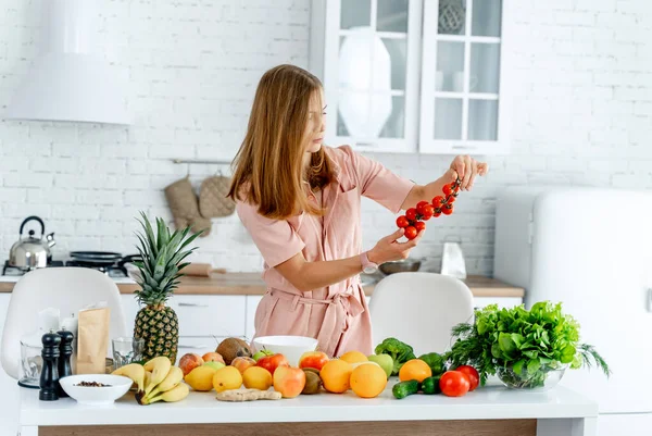 Kvinna i köket redo att laga mat med grönsaker och frukt. Kvinnan tittar på tomater i händerna. Köksbakgrund. Friska livsmedel. Veganer. Gurkväxter. — Stockfoto