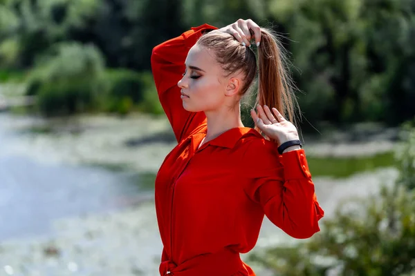 portrait of a beautiful young woman in red, orange dress. Park view. Lake view blurred background. Long hair. Nice makeup. Professional photoshoot. Closeup. Holds hair in hands. Hand near the head.