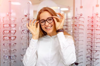 Mutlu kadın optometrist, optik dükkanında arka planda bir sürü bardakla ayakta duruyor. Gözlükleri takın. Görme düzeltmesi. Kadın dioptrik giyiyor. Yakın çekim..