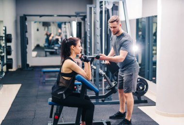 Genç, güzel esmer fitness kızı spor aletleriyle spor salonunda antrenman yapıyor. Antrenör, daha iyi sonuçlar için yeniden uzlaşma sağlıyor.