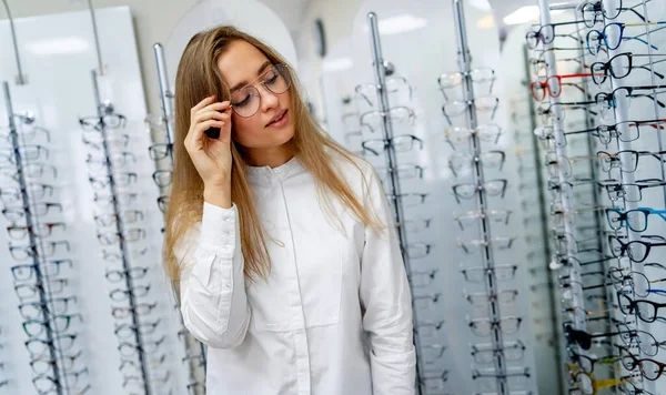 Felice optometrista femminile, ottico è in piedi con molti occhiali in background in negozio ottico. Mettiti con gli occhiali. Correzione della vista. La donna indossa la diottrica. Primo piano . — Foto Stock
