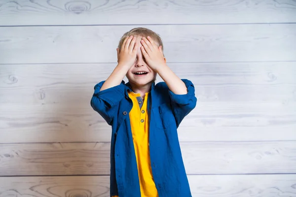 Retrato de um menino bonito em camiseta amarela e casaco de ganga, camisa. Menino de pé sobre um fundo de madeira branco. Rapaz de 5 anos. Fecha olhos com mãos . — Fotografia de Stock