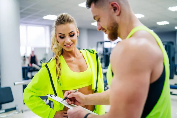 Personal trainer with a folder working with client woman at a gym. Creating a scedule. Wears bright clothes. The woman is in a jacket. Gym background.