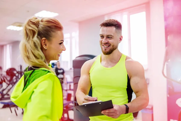 Personal trainer with a folder working with client woman at a gym. Creating a scedule. Wears bright clothes. The woman is in a jacket. Gym background.