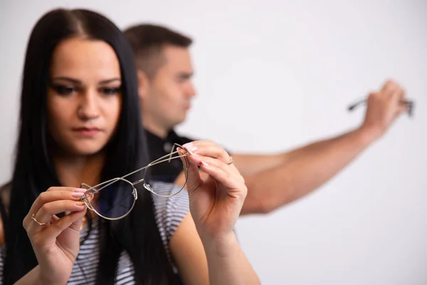 Porträtt av en ung kvinna med glasögon i händerna. En suddig man i bakgrunden. Flickan tittar genom glasögon. Långhåriga brunett vacker flicka och glasögon i händerna. Närbild. — Stockfoto