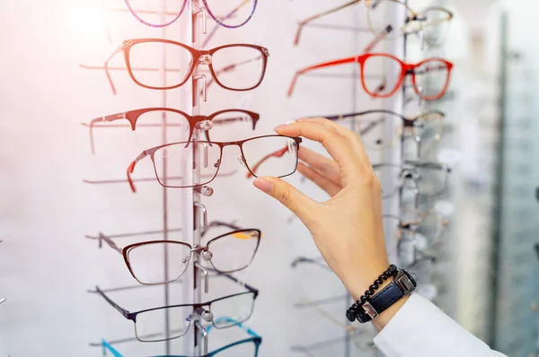 Fila de gafas en una óptica. Tienda de anteojos. Soporte con gafas en la tienda de óptica. La mano de la mujer elige gafas. Corrección de la vista. —  Fotos de Stock