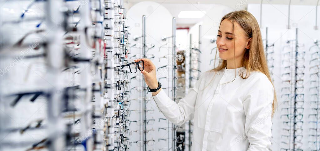 Happy female optometrist, optician is standing with many glasses in background in optical shop. Stand with spectacles. Eyesight correction.