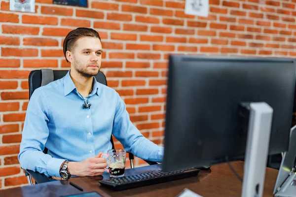 Portrait of young man sitting at desk in the office using computer. Brick wall background. Closeup. Business and management concept. — Stock fotografie