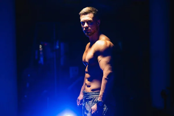 Attractive bodybuilder posing and showing off muscles on dark background. Half turn to the camera. Closeup. Blue light filter. Strong athlete with naked torso. Fitness concept. — Stock Photo, Image