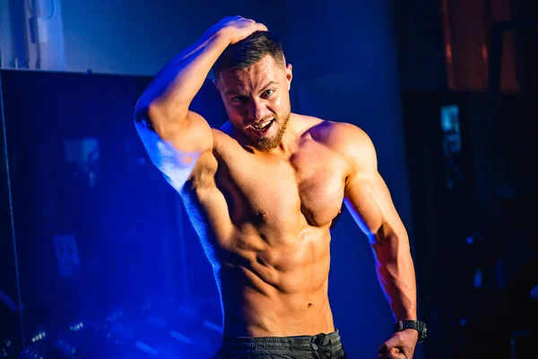 Handsome man with big muscles, posing at the camera in the gym, Black and blue background. Portrait of a smiling bodybuilder. Closeup. — Stock fotografie