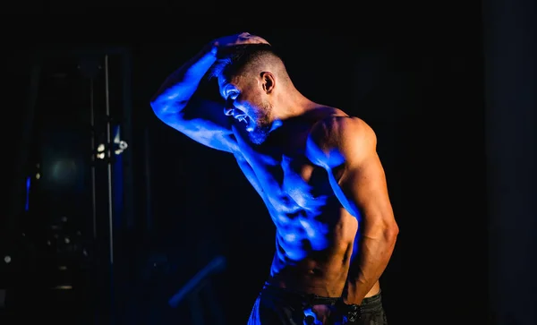 Bodybuilder showing belly and biceps muscles, personal fitness trainer. Strong man flexing muscles. Fitness model posing at camera. Black and blue light background. — Stock fotografie