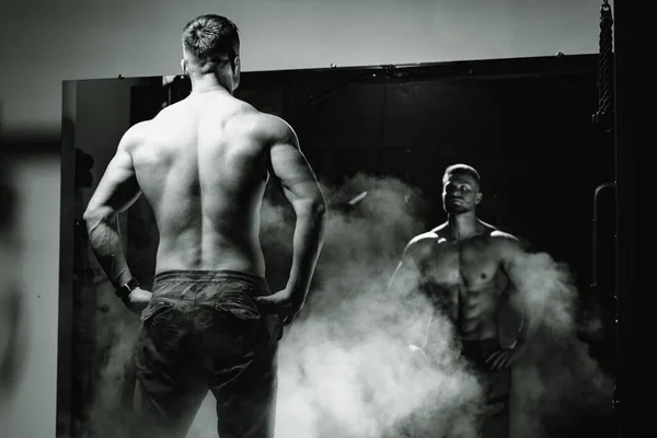 An athletic man standing and posing in front of the mirror. View from the back. Black and white photo. Smoke and power. Fitness concept.