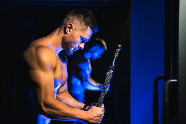 Handsome brutal sexy strong bodybuilder athletic fitness man pumping up muscles and doing workout with trx. Bodybuilding concept. Black and blue light background. View from the side closeup. — Stockfoto