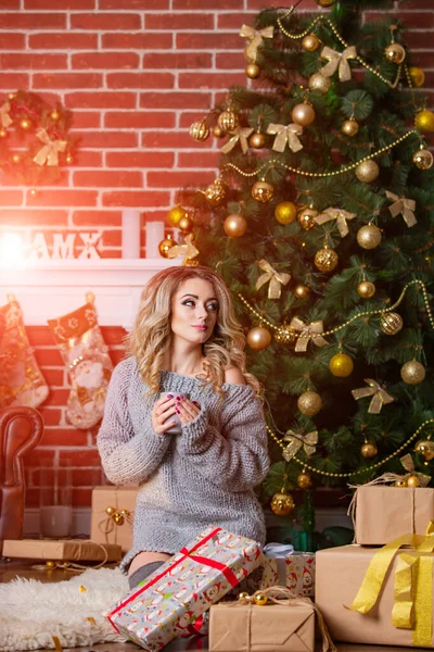 Young beautiful blonde woman is sitting under the Christmas tree. New Year decorations. Brick wall background. Girl in grey knitted sweater with presents. Portrait of a nice girl. — 스톡 사진