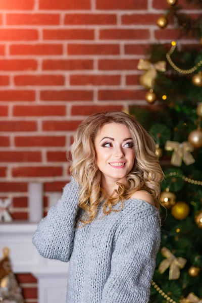 Young beautiful blonde woman near the Christmas tree. New Year decorations. Brick wall background. Feast preparations. Girl in grey knitted sweater in a blurred background. — 스톡 사진