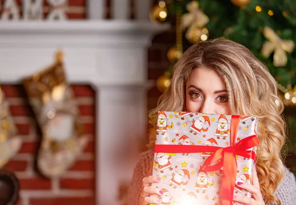 Girl in a sweater near the Christmas tree with a gift near face, New Year Christmas tree. Young woman in anticipation of New Year. Blurred background. Long haired blonde. — 스톡 사진