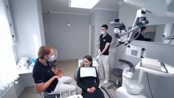 歯の椅子に座っている女性 現代の歯科事務所の背景にある魅力的な女性の訪問の速水科医 マスクの歯科医はクリニックの患者と話します — ストック動画
