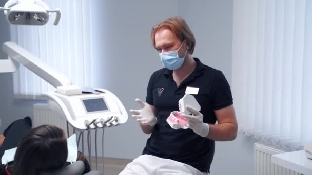 熟练的医生显示出假牙 英俊的牙医坐在一个现代化的牙科诊所的椅子上与病人交谈 牙科医生假牙咬牙切齿 牙科治疗概念 — 图库视频影像
