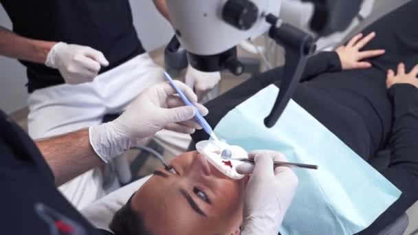 若い女性は歯の椅子に横たわっていた 歯医者は歯医者で患者の歯を治療する 組織学者は顕微鏡を通して歯を調べる — ストック動画