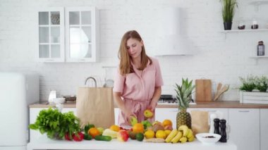 Sağlıklı vitaminleri boşaltan mutlu kadın. Evdeki mutfakta genç bir bayan. Masanın yanında bir sürü taze meyve ve sebzeyle gülümseyen ev hanımı. Diyet yaşam tarzı.