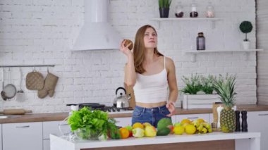 Alışverişten sonra hindistan cevizli güzel bir kadın. Elinde taze meyve tutan ve mutfak arka planında gülümseyen genç bir bayan. Masada taze meyve ve sebze var. Sağlıklı gıda konsepti.