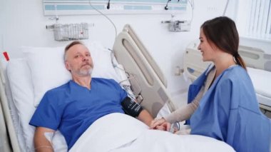 Gülen kadın hasta hastayla konuşuyor. Genç bayan ziyaretçi hastane yatağında yaşlı adamın yanında oturuyor. Ameliyathanede çağdaş ekipmanlar var..