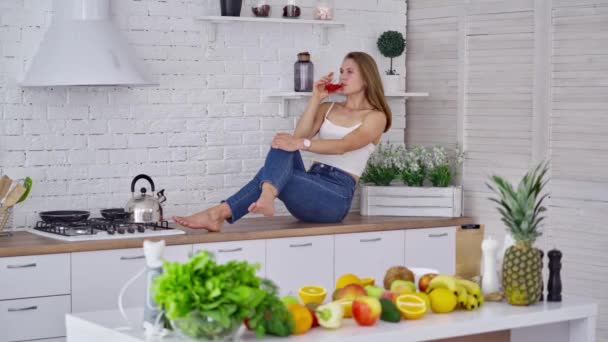 厨房桌上漂亮的模特儿喝果汁 穿着牛仔裤的年轻妇女坐在桌子上 在家里喝果汁 健康食物及饮品的概念 — 图库视频影像