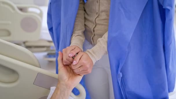 病気の患者をサポートするクローズアップ女性の手 女性の訪問者は病院の部屋で父親の手を握っている シニア男性患者はクリニックでベッドに横たわっています — ストック動画