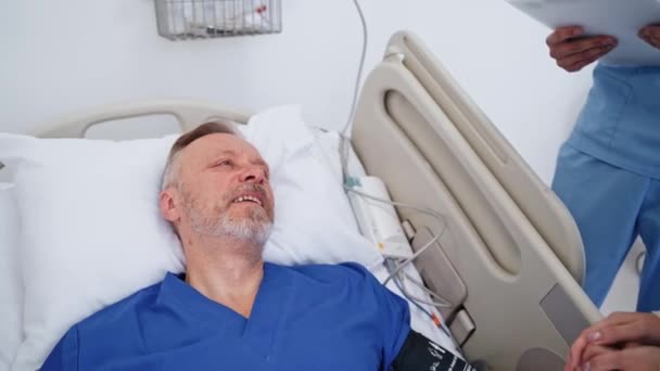 老年人躺在医院的床上 生病的男性病人在诊所里与来访者愉快地交谈 保健概念 — 图库视频影像