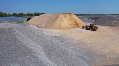 Dışarıda ezilmiş taş ve kum yığınları var. Buldozer güneşli bir günde asfalt fabrikasında çalışıyor. Hava görünümü.