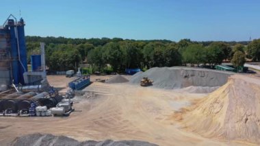 Yazın doğa ile çevrili sanayi bölgesi. Asfalt bitkisi, kum ve moloz yığınları. Kazı araçları çakıl taşır. Hava görünümü.