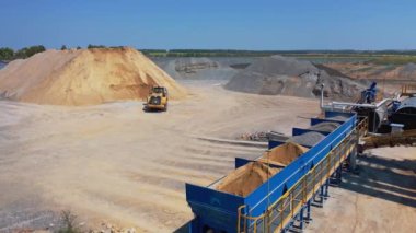 Buldozer kovada kum taşır. Asfalt bitkisi ezilmiş taş ve çakıl yığınlarıyla çevrili. Modern bir fabrikanın sanayi bölgesi..
