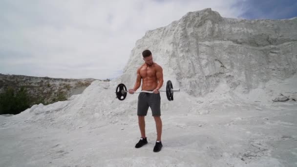 屋外でのハンサムなアスリートのトレーニング 夏には白い山の背景に筋肉質の体を持つ強い無気力な男が鐘を持ち上げています スローモーション — ストック動画